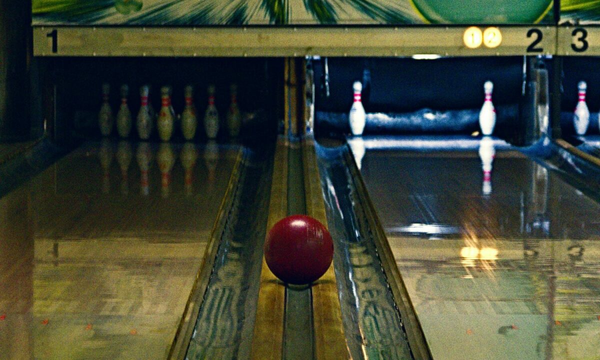 A maroon bowling ball returning to the bowler between two lanes in an old bowling alley. One lane is lit up showing a 7 and 10 split.